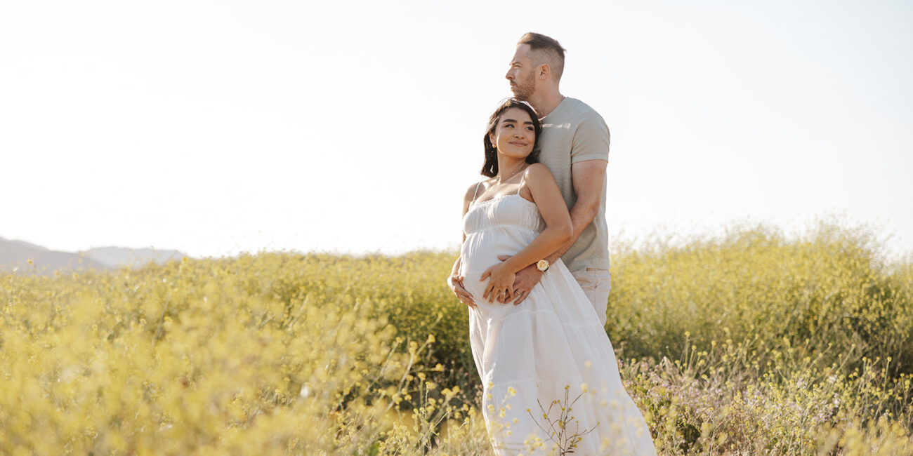 A pregnant woman in a white dress stands in a field with her husband behind her, in California - portrait photography by Leah Marie Photography