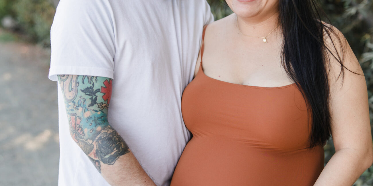 A smiling pregnant woman in a rust-colored dress stands outdoors with husband, in California - lifestyle photography by Leah Marie Photography