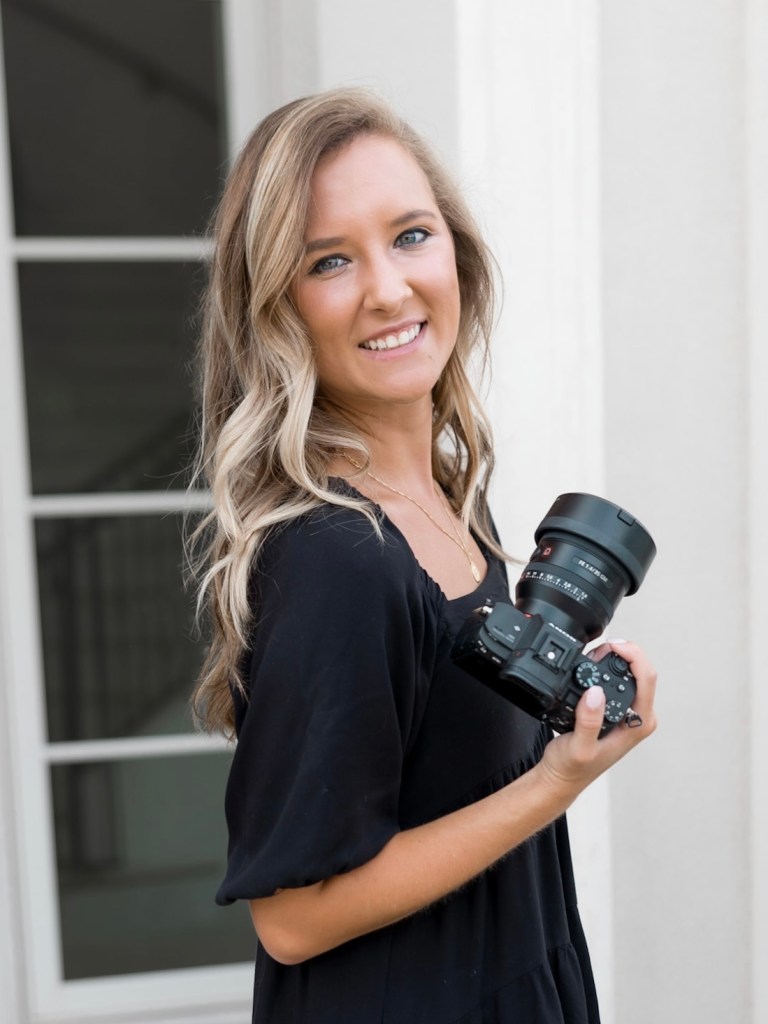 A woman with long, wavy blonde hair wearing a black dress smiles at the camera while holding a large, professional camera in her right hand. She stands in front of a white wall and window. Leah Marie Photography - southern california