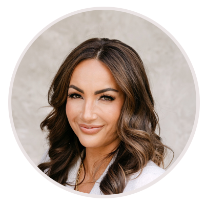A woman with long, wavy brown hair and natural makeup smiles gently at the camera, wearing a white top against a light, neutral background. Leah Marie Photography - southern california