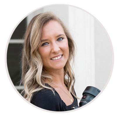 A woman with long, wavy blonde hair smiles at the camera while holding a camera lens. She is wearing a black top and standing in front of a light-colored building. Leah Marie Photography - southern california