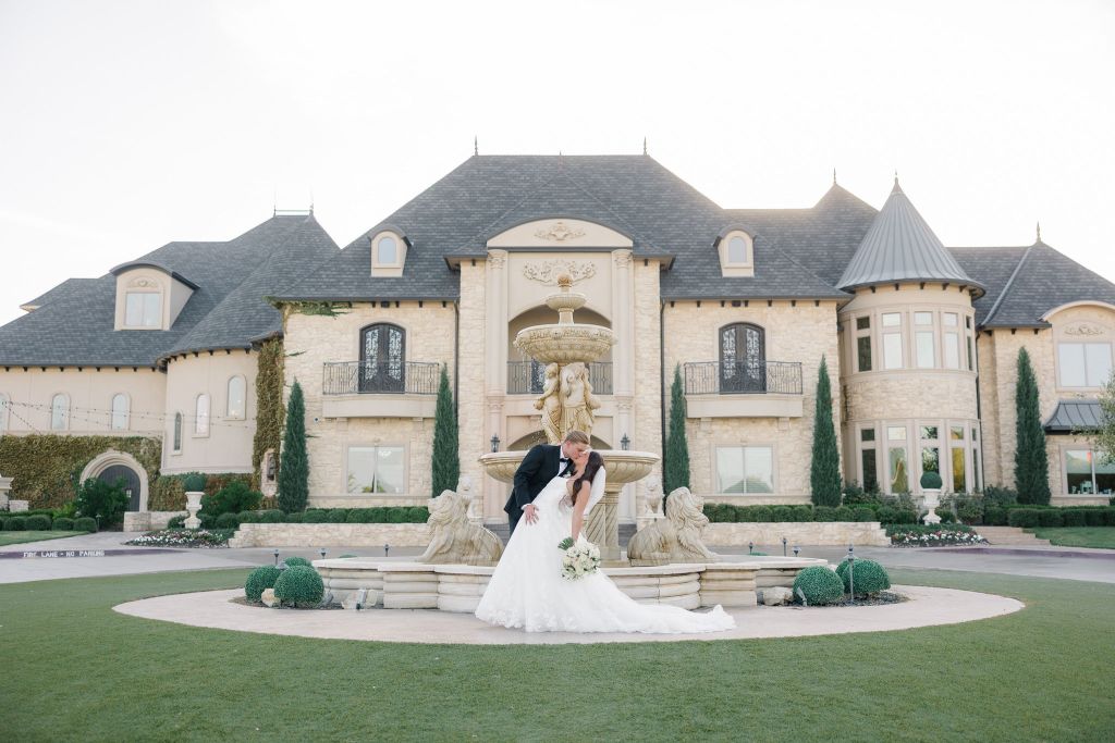 A bride and groom kiss in front of a large, elegant mansion with a central fountain and manicured landscaping. The bride wears a white gown and holds a bouquet, while the groom is in a black suit. Leah Marie Photography - southern california