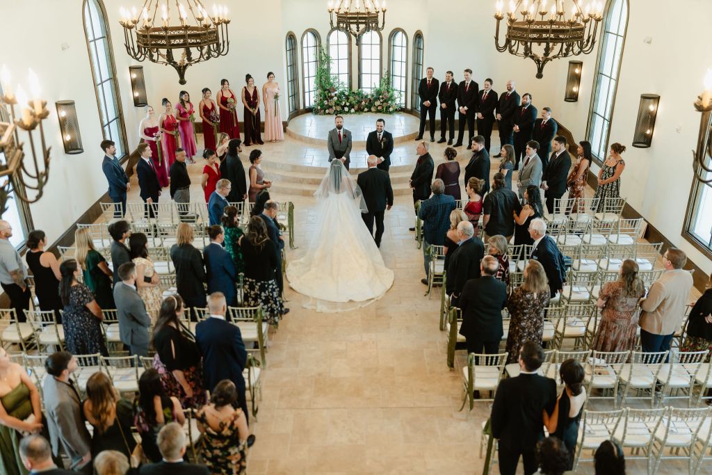 A bride in a white gown walks down the aisle with an escort toward the groom and officiant at the altar in a bright chapel, surrounded by seated guests and a wedding party in formal attire. Leah Marie Photography - southern california