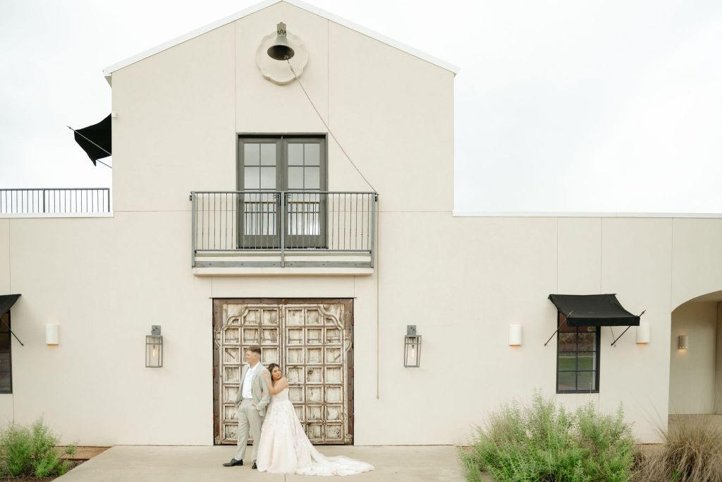 A bride and groom stand in front of a large, light-colored building with a balcony and a decorative wooden door. The bride wears a strapless gown and the groom is in a light suit. Green shrubs frame the foreground. Leah Marie Photography - southern california