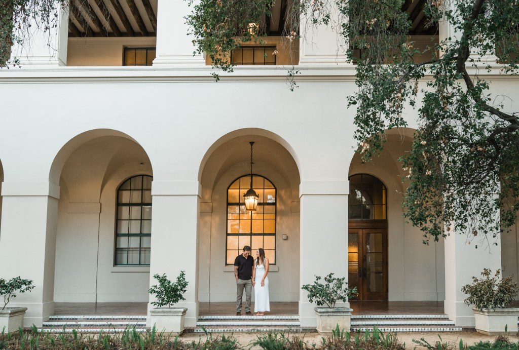 A couple stands together under an archway of a large, white building with tall columns and arched windows. Green trees and plants frame the foreground. Leah Marie Photography - southern california